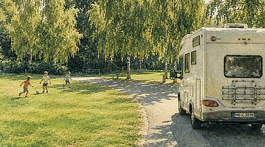 Wohnmobil auf dem Weg unter Birken, im Hintergrund spielen Kinder Fußball auf der Wiese.